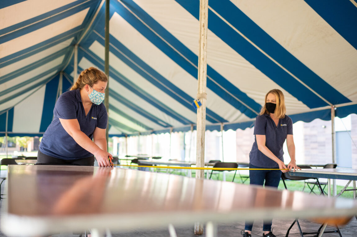 Photo of Jennifer Pomerantz (left) and Elaine Hanke of Michigan Conference & Event Services measuring the distance between tables inside a tent on North Campus.