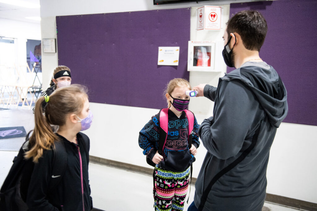 Three young kids get temperature checked by adult.