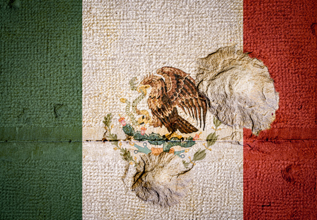 Bullet holes a rough wall with the flag of Mexico. Concept of violence in Mexico and US-Mexico Border Militarization and brutality. Image credit: iStock