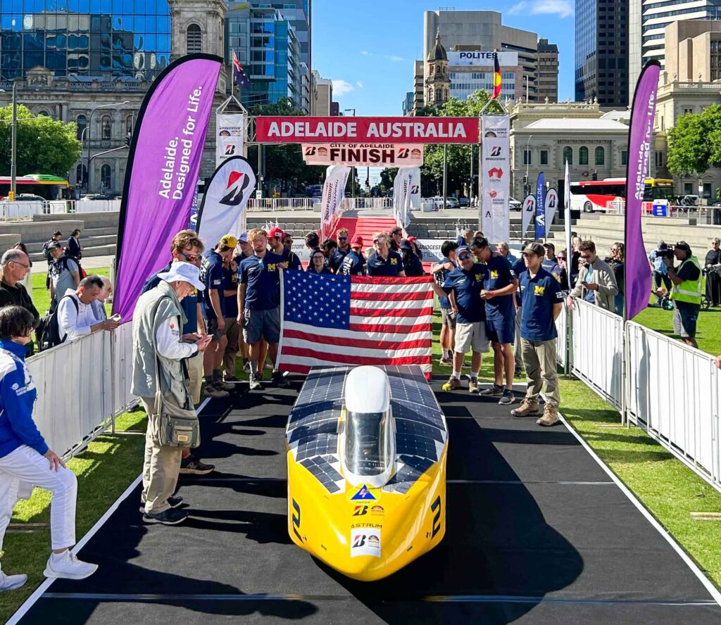 The Michigan solar car team stands behind their yellow car, Astrum, at the ceremonial finish line in Adelaide, Australia. Tall buildings are visible in the background, and a red banner reads "Adelaide Australia" over the. The entire team is wearing maize and blue and are holding up the American flag.