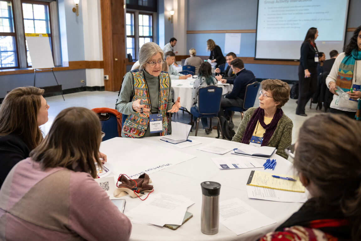 A photo of a woman standing at a round table talking to several people