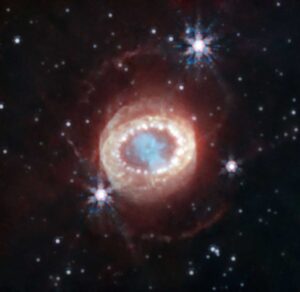 On a black backdrop, dim or distant stars glow white, with three nearer or brighter stars producing hexagonal diffraction patterns like rainbow snowflakes. In the middle of the frame, the supernova remnant resembles a reptilian eye, with a teal cloud in the center ringed by bright white dots that are the clumps of hydrogen lit up by the shockwave from the supernova. A pale haze surrounds the ring of dots, with other less distinct dots inside. A near-infrared image of the remnant left behind by supernova 1987A, taken by the James Webb Space Telescope. The hydrogen clumps known as the "string of pearls" appear as a ring of white dots around the teal center of the stellar remnant, still shining brightly due to the energy imparted by the supernova shockwave. The number of clumps is consistent with the Crow instability having caused them to form. Image credit:  NASA, ESA, CSA, M. Matsuura (Cardiff University), R. Arendt (NASA’s Goddard Spaceflight Center & University of Maryland, Baltimore County), C. Fransson (Stockholm University), J. Larsson (KTH Royal Institute of Technology), A. Pagan (STScI)