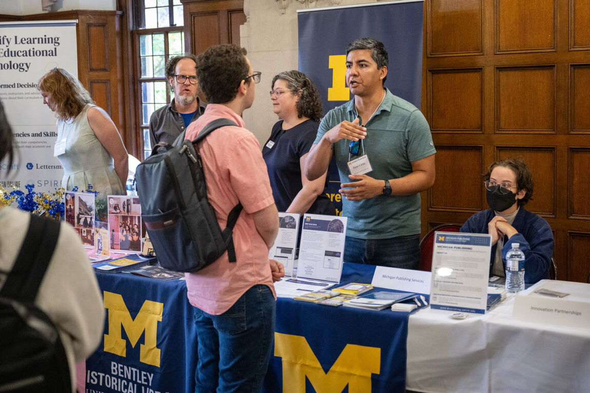A photo of a man talking to a person while others gather around long tables