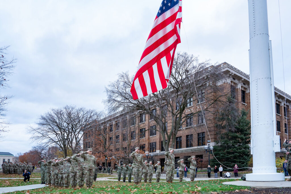 Photo of rows of ROTC students in uniform saluting as the American flag is raised over the Diag.