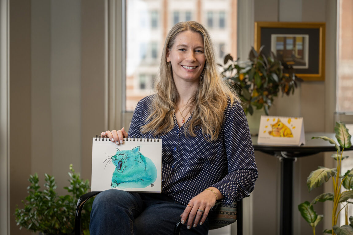 A woman sitting in a chair holding a watercolor painting of a cat she made