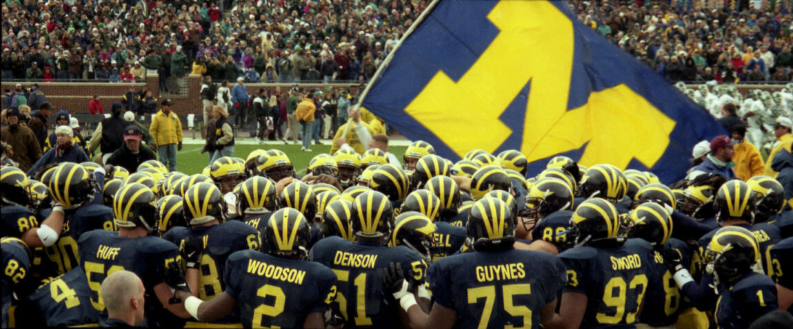 A photo of the U-M football team huddling with a Block M flag in the background
