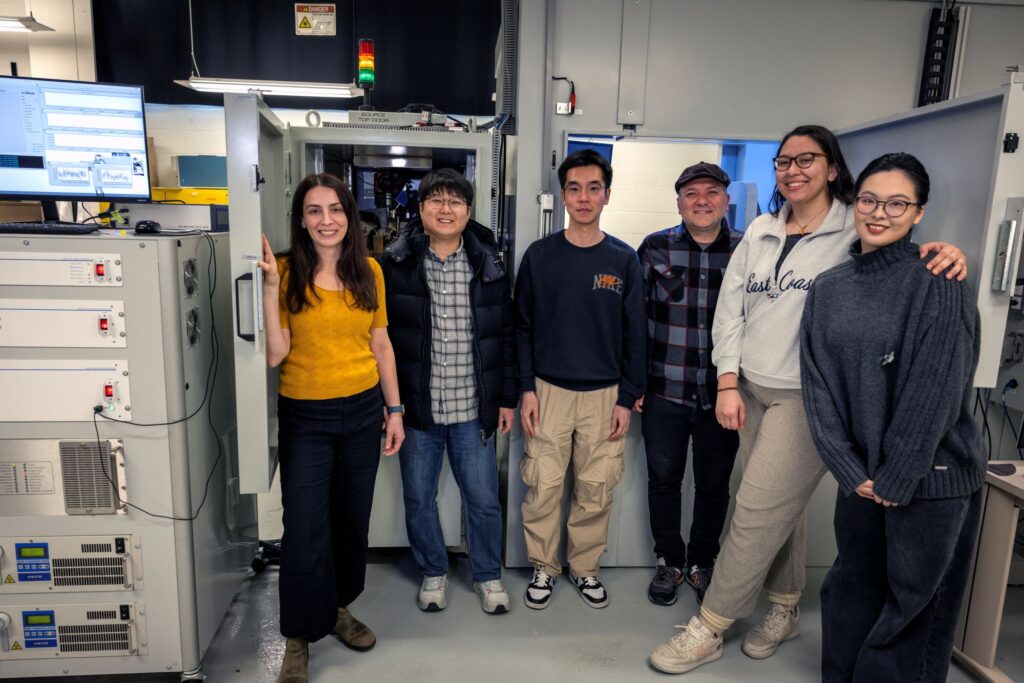 Six people stand side-by-side in a laboratory in front of a large device that resembles a 5-foot-tall refrigerator with microscope equipment inside the top drawer.
