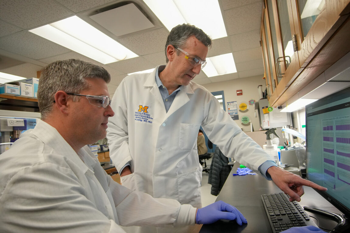 Two men in lab coats pointing at a computer screen