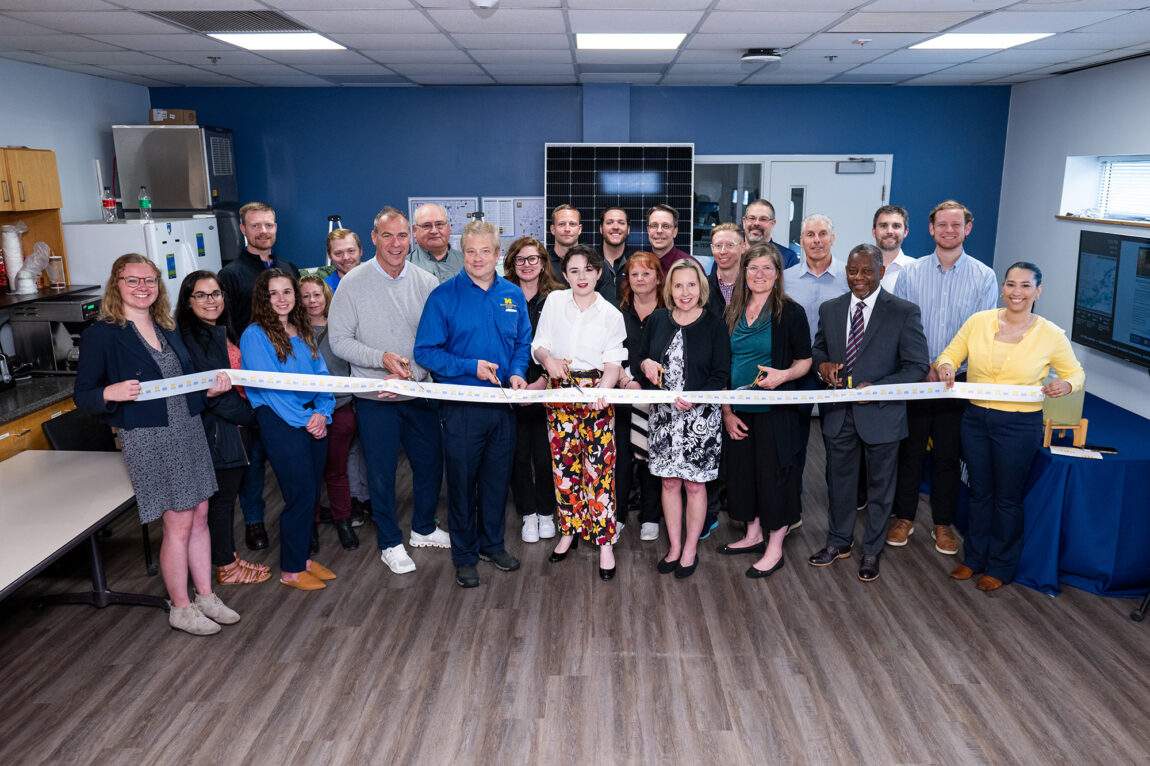 A group of people standing with a white ribbon, and several of them are pretending to cut the ribbon with scissors