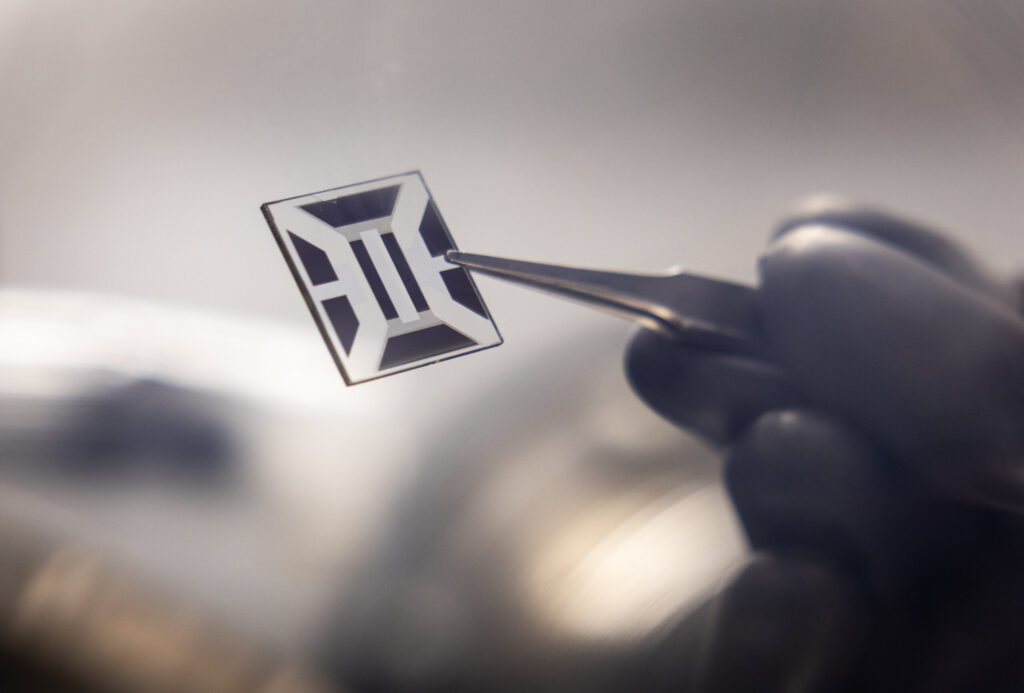 A transparent square, a couple of centimeters to a side, with black marks on it, is held in a pair of tweezers.


