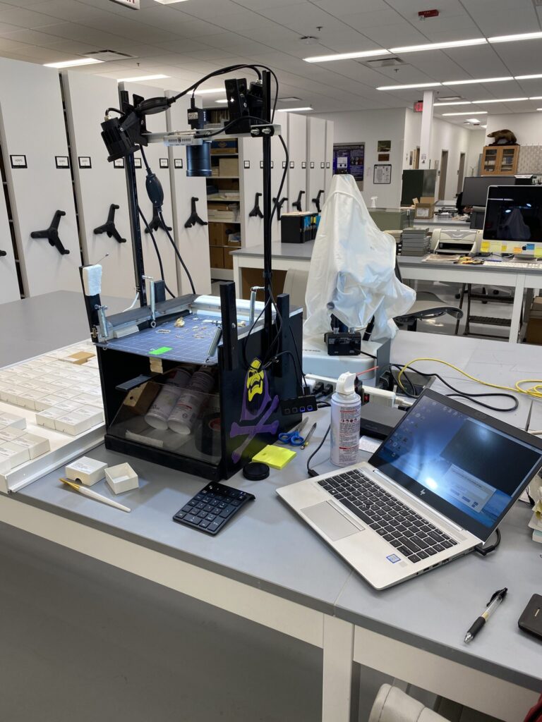 The Skelevsion system is set up on a table, in front of rows of specimen cabinets, and consists of a box-like platform where samples can be observed. A camera is mounted above the platform, looking down at the specimens, and the system is connected to a laptop computer. 