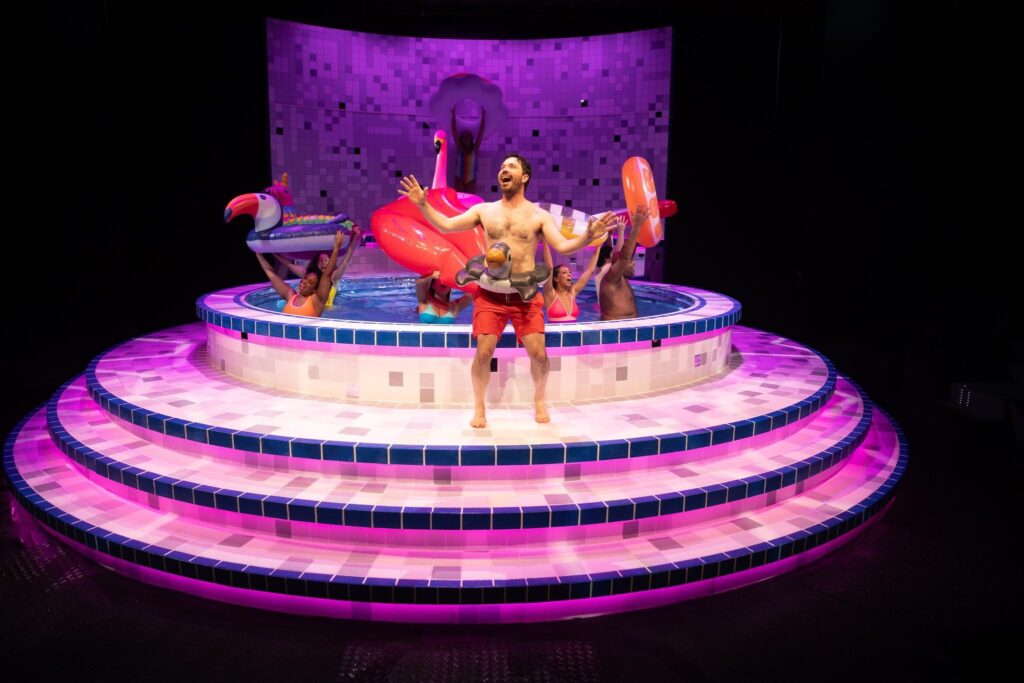 A man in swimtrunks and wearing a floatation device around his waist stands near a pool on a stage