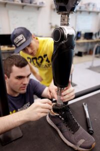 Seelhoff appears to adjust the athletic shoe covering the foot of the prosthesis. Best leans in to steady the prosthesis with a hand on the metal post that links the foot to the black, calf-shaped box housing the motor and electronics.