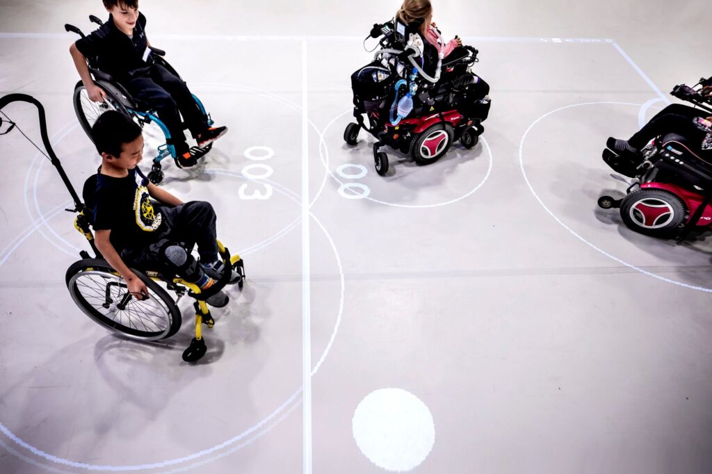 Two children in manual wheelchairs play against peers in power chairs, the ball appearing as a white spot in an open space. The lines from the projector bound a rectangle around them and the center line between them as well as arcs marking the goals.