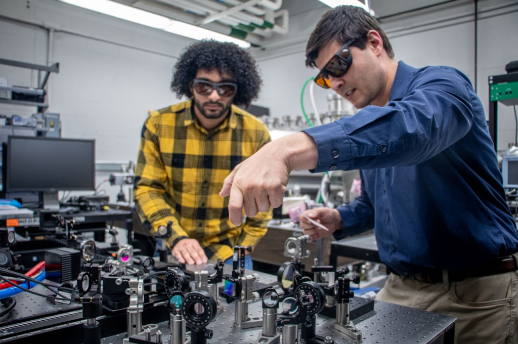 Two men in dark protective glasses work at an optical table, a black surface with optical components like lenses and reflectors secured to it.