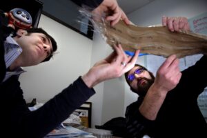 Two people examine a piece of brown fabric with photonic fibers in a room. The photonic fibers are visible as shiny strands protruding from one end.