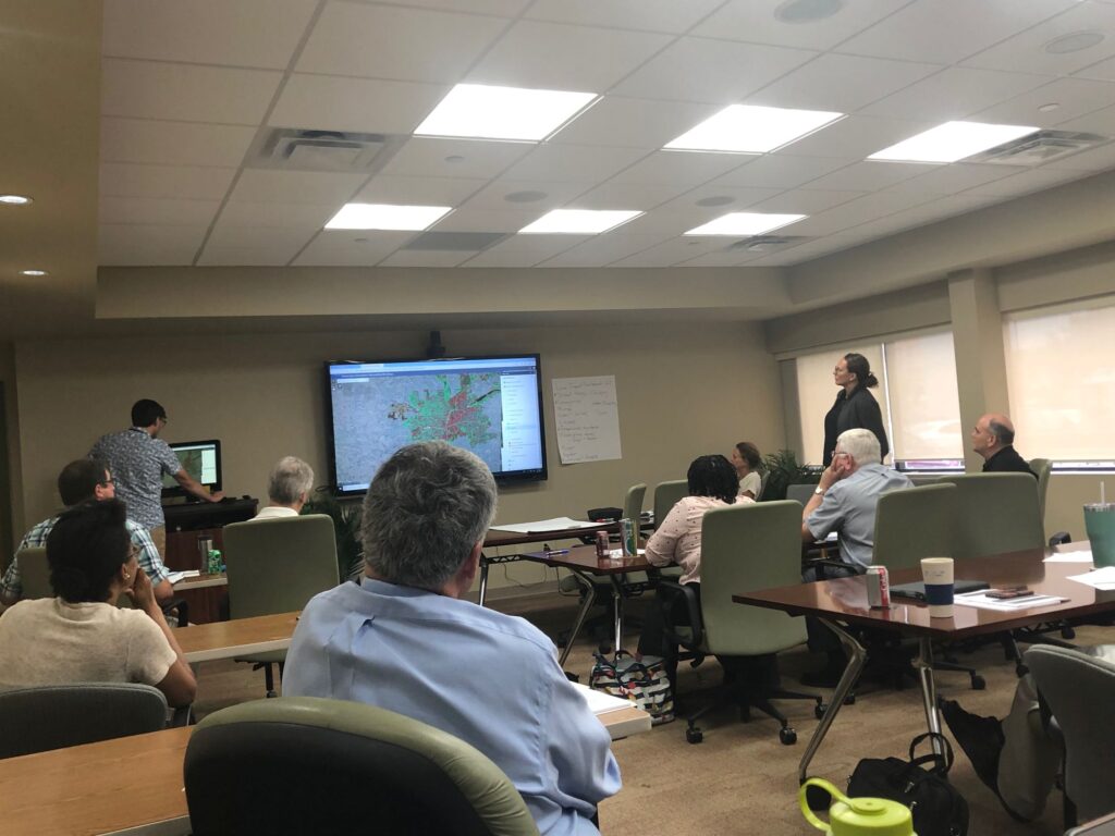 Several people sit in a conference room looking toward a large screen at the front of the room showing a map highlighted with red and green splotches.
