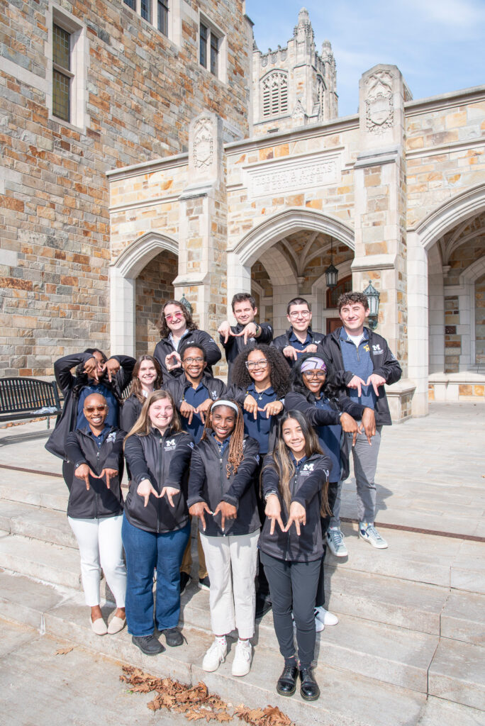 Thirteen people standing on steps in three rows pose for a photo while making "M" shapes with their hands