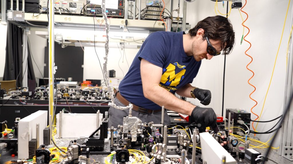 A man in black gloves and laser safety glasses stands at a crowded optical table with various elements attached, some with wires running to them.