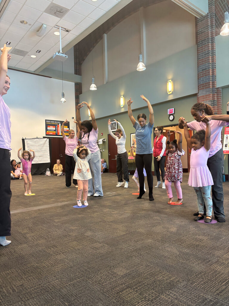 Young women and children gather in a room in a ballet pose