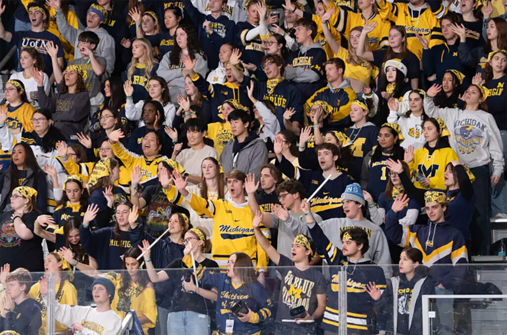 Fans dressed mostly in maize and hockey gear cheer at an indoor ice hockey game
