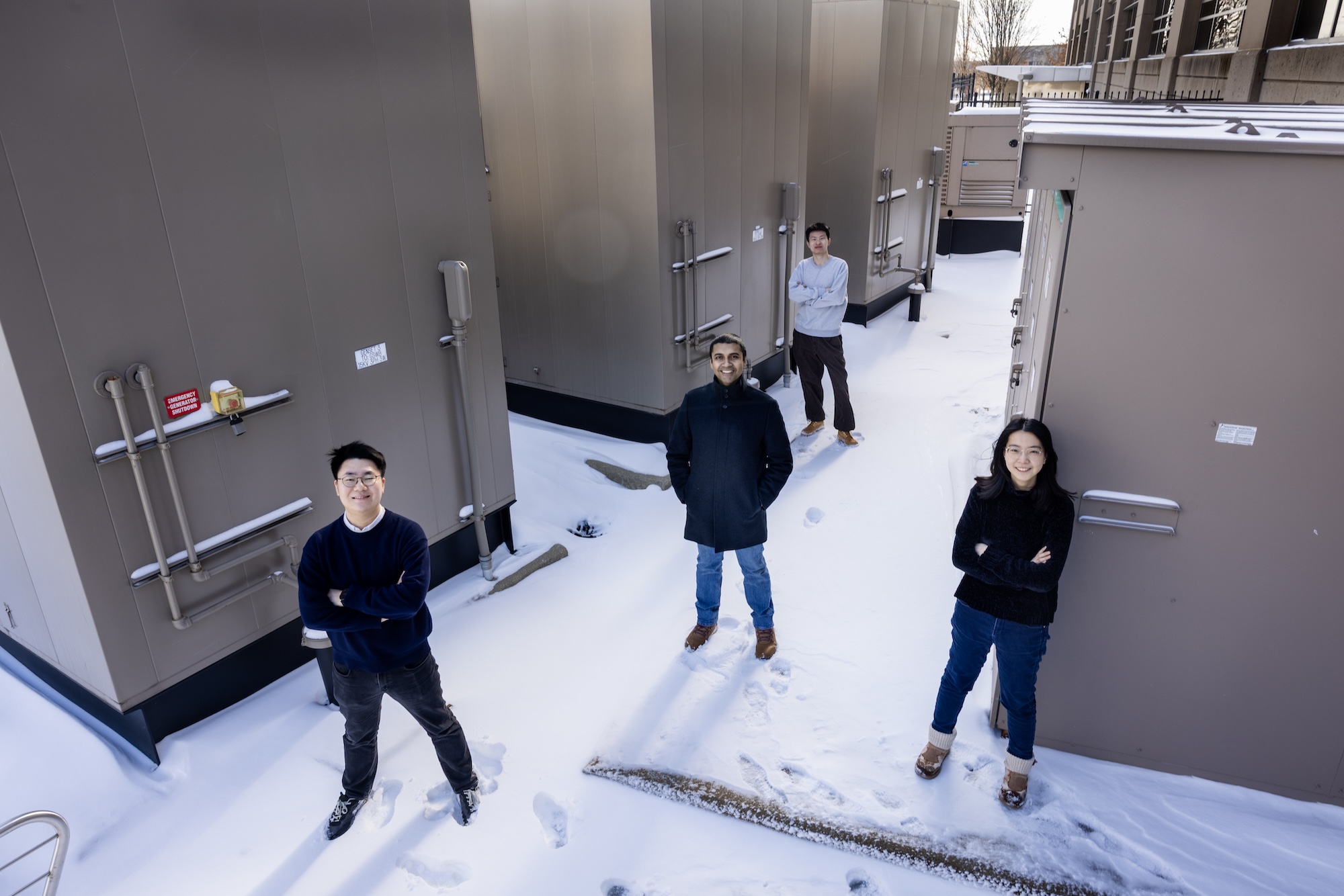 Three men and a woman stand in a triangle formation among generators outside a computing center. The generators are inside tan sheds that are two or three times taller than the researchers. There is snow on the ground.