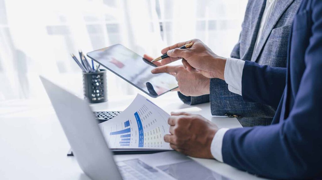 Tight crop of the hands of two people wearing suits and a laptop discussing financial documents. Image credit: Adobe Stock