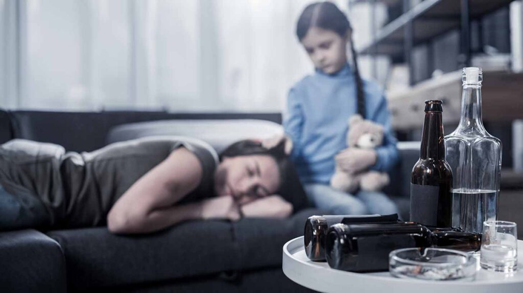 A child watches over her mother passed out on the couch with alcohol and ashtrays in the foreground. Image credit: Adobe Stock