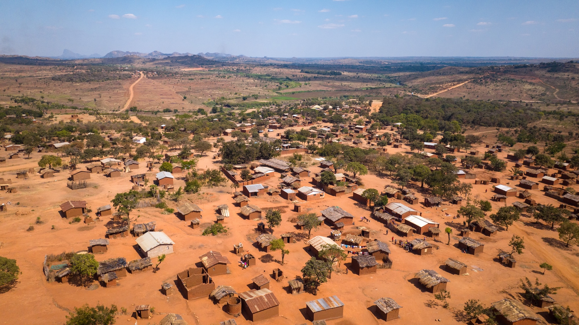 An aerial view shows several small brown buildings in a Malawian village with a savannah stretching out into the horizon. Some of the buildings have rusted metal roofs, while others are covered with thatching. Some have no covering.