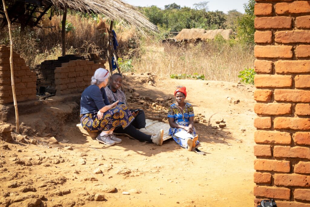 Pam Jagger sits in the shade of a brick structure with a straw-thatched roof speaking with a Malawian man and woman.