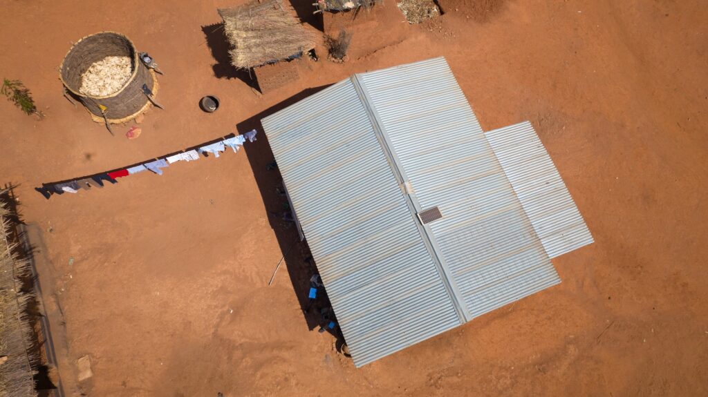 An aerial photograph shows a small solar panel resting on top of a metal roof on a building in Malawi. Outside the building are smaller structures with thatched roofs and clothes drying on a line.