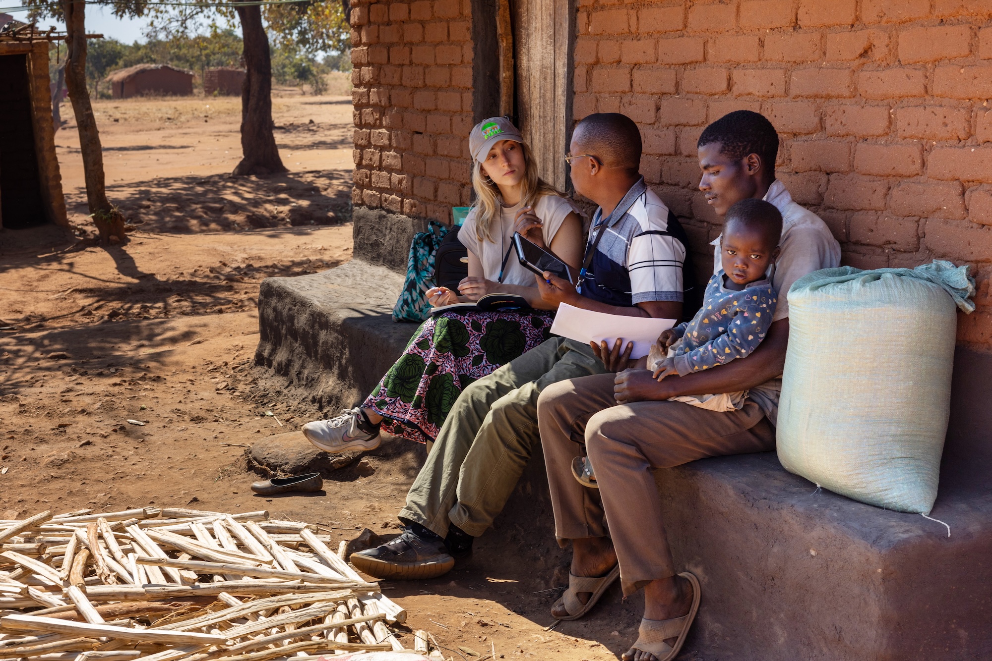 Andrea Mahieu takes notes while speaking with two Malawian men and a child in the shade next to a brick building.