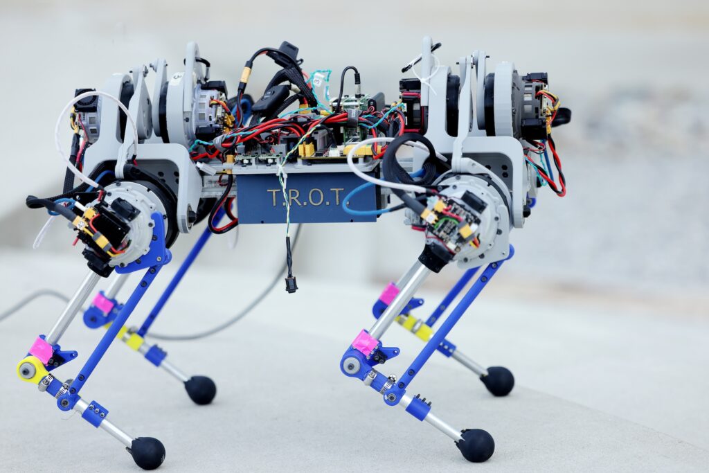 The robot has four legs with backward-facing knees. Each leg has three actuators, controlling hip ab/adduction, hip flexion/extension, and knee flexion/extension. Exposed wires link up to the motors. T.R.O.T is emblazoned on the case around the belly of the robot.
