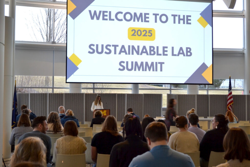 A person stands at a podium and speaks while many others sit facing her with a large sign above that says "Welcome to the 2025 sustainable lab summit"