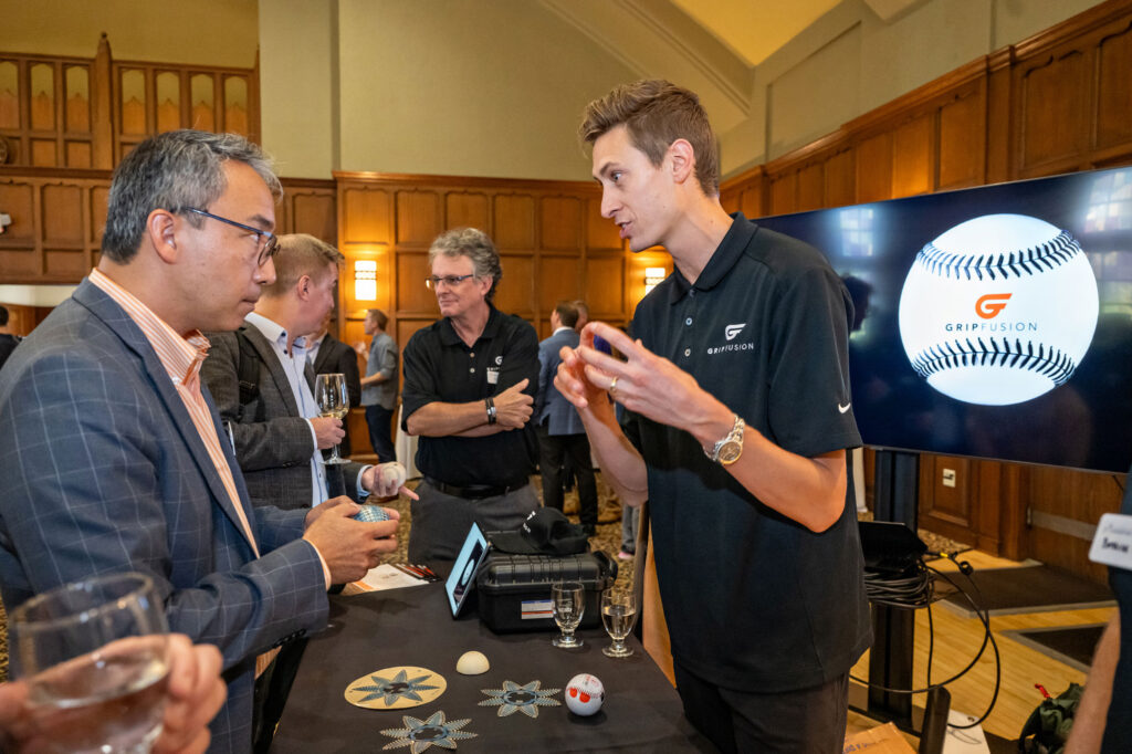 Two men standing at a table discussing the invention of a new type of baseball that measures grip.