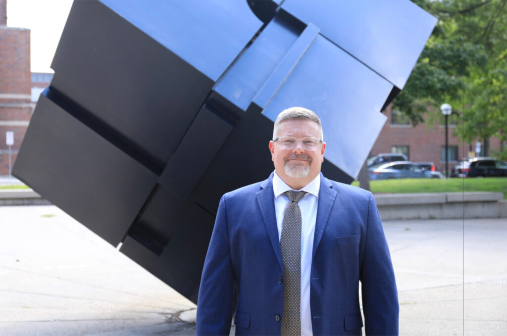 A man in a suit stands in front of a cube structure and smiles