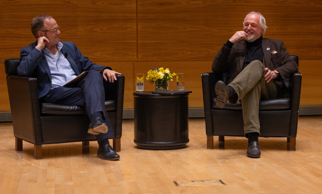 Two men sitting in chairs on a wooden stage speaking to each other