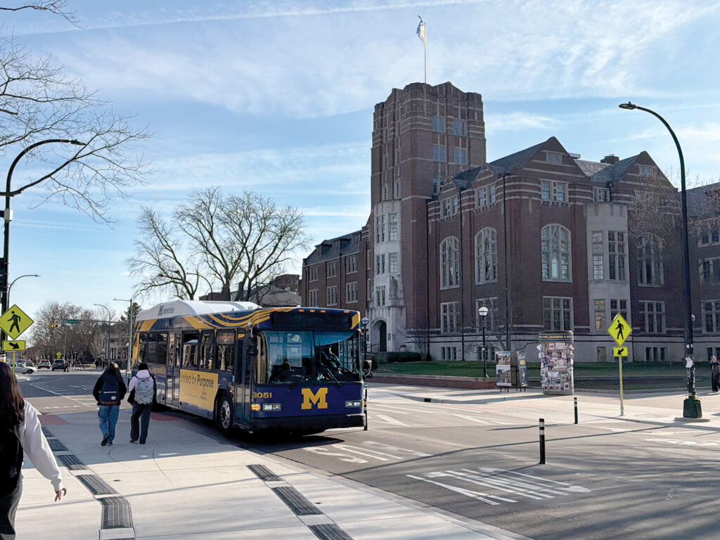 A bus drives along a bus-only lane while pedestrians walk on a sidewalk and a large brick building sits in the background
