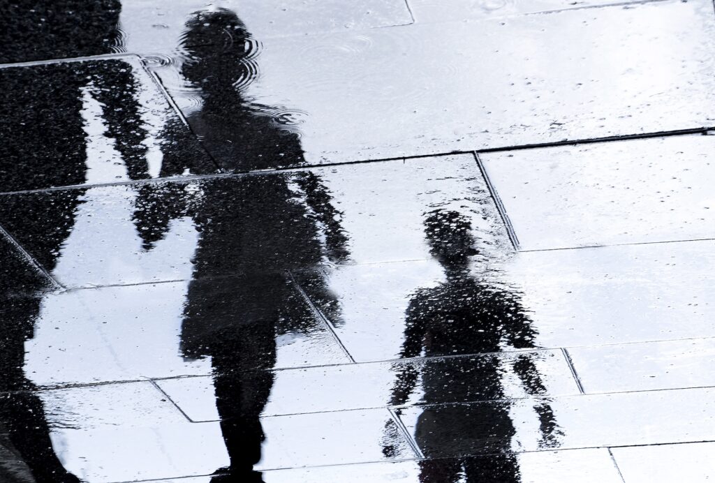Blurry street reflection showing the shadowy silhouettes of siblings walking alongside a parent, captured on a city sidewalk. Image credit: iStock
