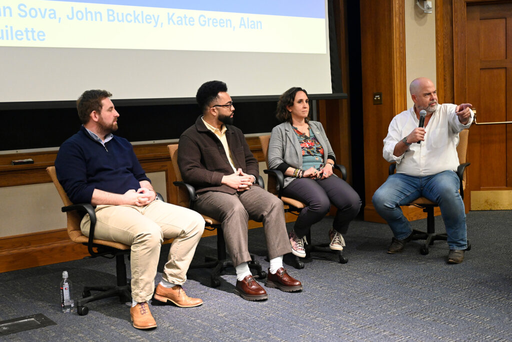 Four people sit in chairs next to each other with one pointing to a crowd with a microphone in his hand