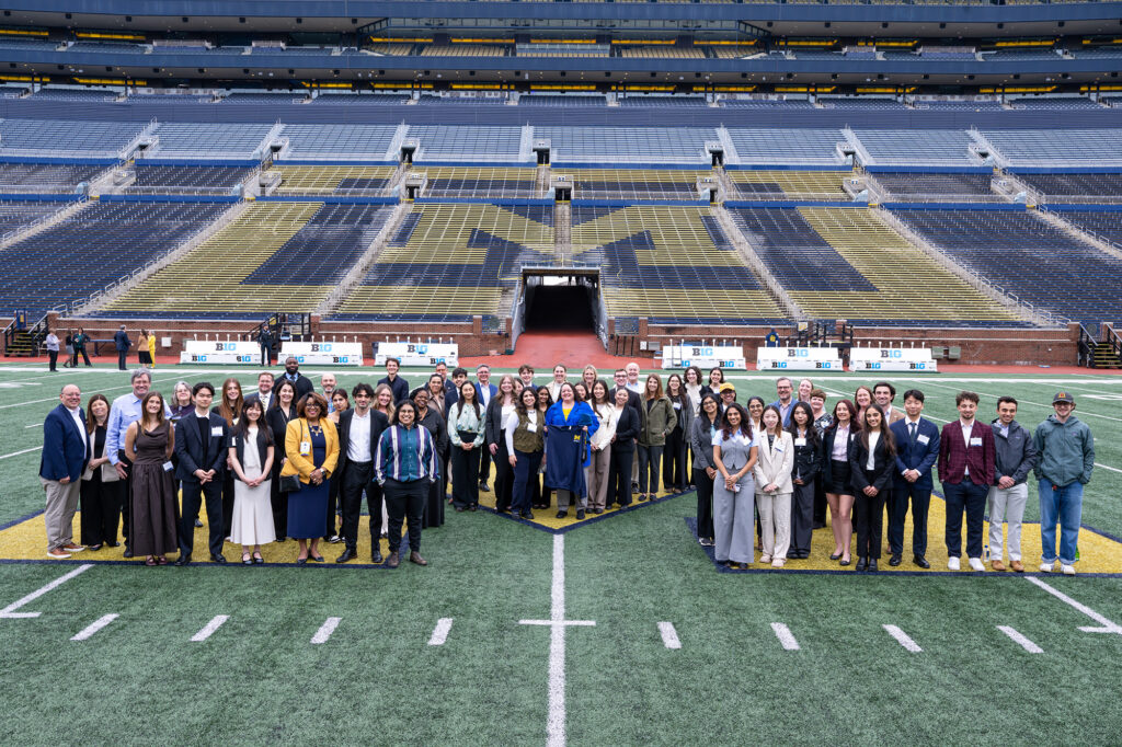 Dozens of people stand on a large block M in the middle of a football field