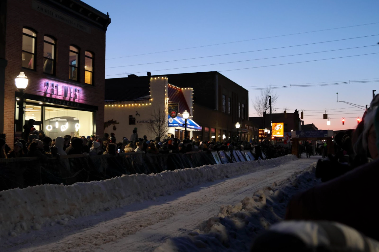 A picture taken up the road of the starting line for the UP 200 race in Marquette, Michigan. Spectators line both sides of the street in front of lighted storefronts. A flat path in the snow has been cleared for the dogsleds, with knee-high snowbanks on both sides of the road.