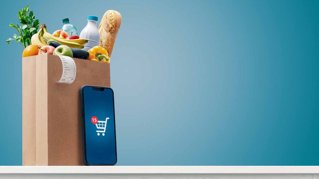 A smart phone displaying a shopping cart propped against a bag of groceries. Image credit: Adobe Stock