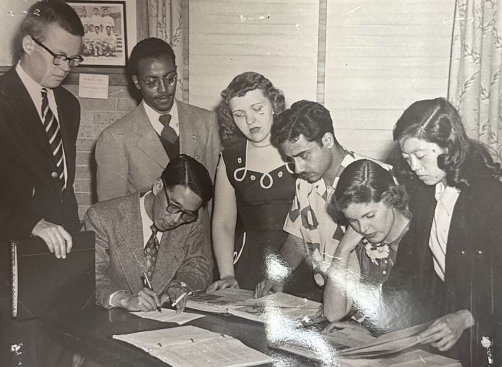 A black and white photo of several people sitting or standing around a table looking at papers