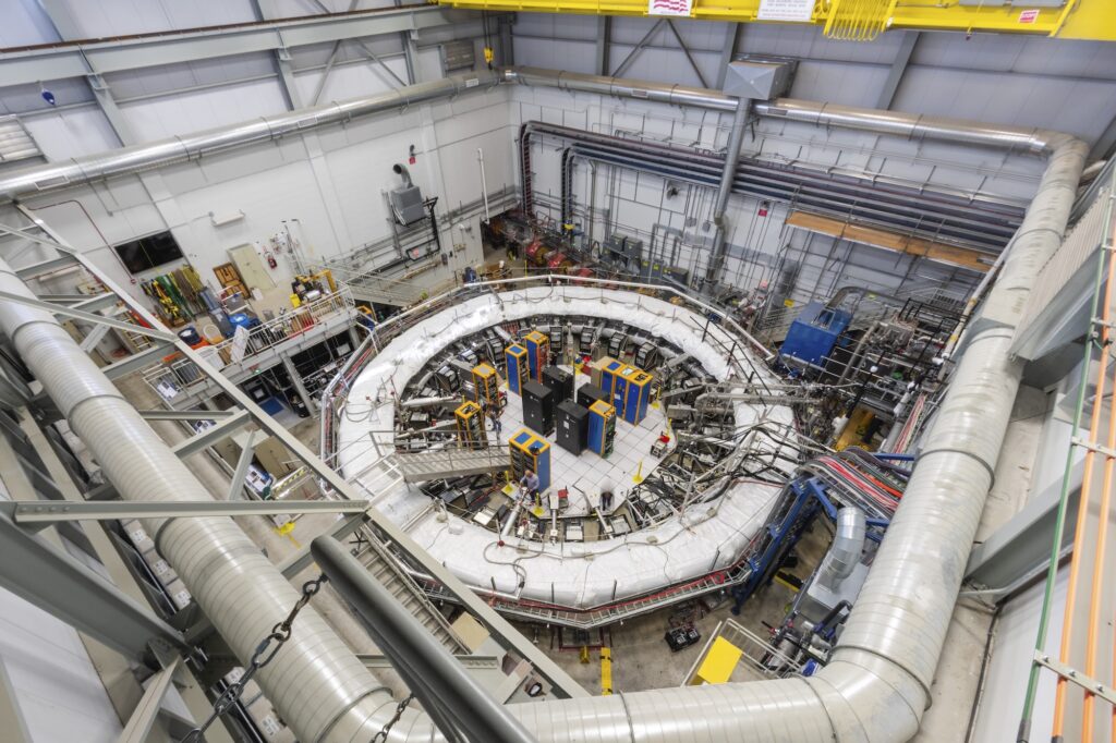 A 50-foot wide white ring sits in the center of a large room, enclosing black, blue and yellow equipment racks. The ring is surrounded by ducts, wiring and the occasional staircase.